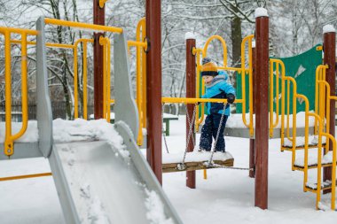 Kar yağışlı kış gününde oyun parkında eğlenen sevimli küçük çocuk. Karda oynayan, kalın giysiler giymiş şirin bir çocuk. Çocuklu aileler için kış etkinlikleri.