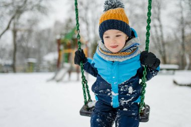 Kar yağışlı kış gününde oyun parkında eğlenen sevimli küçük çocuk. Karda oynayan, kalın giysiler giymiş şirin bir çocuk. Çocuklu aileler için kış etkinlikleri.