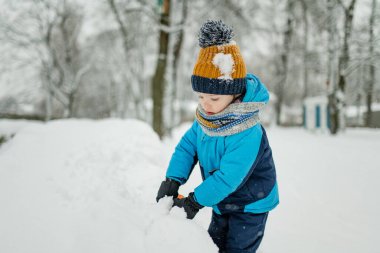 Sevimli küçük çocuk karlı kış gününde eğleniyor. Karda oynayan, kalın giysiler giymiş şirin bir çocuk. Çocuklu aileler için kış etkinlikleri.