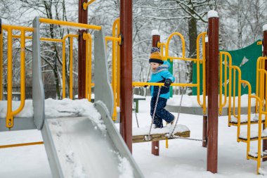 Kar yağışlı kış gününde oyun parkında eğlenen sevimli küçük çocuk. Karda oynayan, kalın giysiler giymiş şirin bir çocuk. Çocuklu aileler için kış etkinlikleri.
