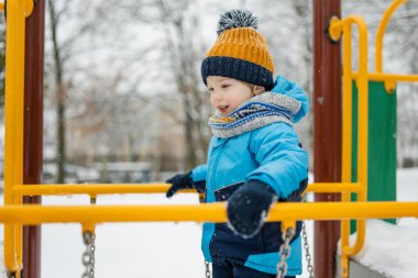 Kar yağışlı kış gününde oyun parkında eğlenen sevimli küçük çocuk. Karda oynayan, kalın giysiler giymiş şirin bir çocuk. Çocuklu aileler için kış etkinlikleri.