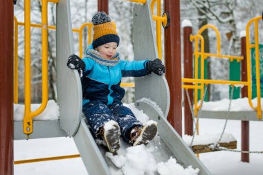 Kar yağışlı kış gününde oyun parkında eğlenen sevimli küçük çocuk. Karda oynayan, kalın giysiler giymiş şirin bir çocuk. Çocuklu aileler için kış etkinlikleri.