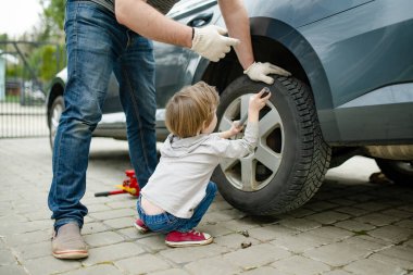 Küçük tatlı çocuk, babasının arka bahçesinde araba tekerleklerini değiştirmesine yardım ediyor. Baba küçük oğluna alet kullanmayı öğretiyor. Küçük bir çocuğun aktif ebeveyni.