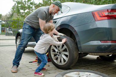 Küçük tatlı çocuk, babasının arka bahçesinde araba tekerleklerini değiştirmesine yardım ediyor. Baba küçük oğluna alet kullanmayı öğretiyor. Küçük bir çocuğun aktif ebeveyni.