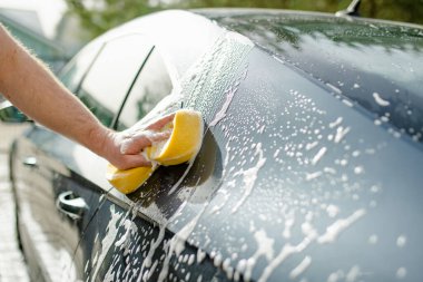 Genç bir adam arabasını yıkamak için sünger kullanıyor. Erkek sürücü, yüksek basınçlı su jeti ile araba yıkıyor. Arka bahçede bir aracı temizlemek..