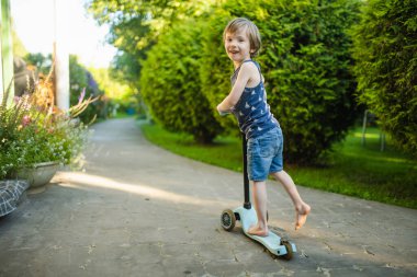 Güneşli bir yaz akşamında arka bahçede scooterını süren sevimli küçük bir çocuk. Paten süren küçük bir çocuk. Çocuklar için aktif eğlence ve açık hava sporları.