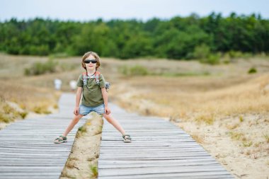 Güneş gözlüğü takan neşeli bir çocuk ve Litvanya 'daki Curonian Spit' in tahta kaldırımında neşeli bir şekilde koşan bir sırt çantası, güneşli bir açık hava macerasının tadını çıkarıyor..