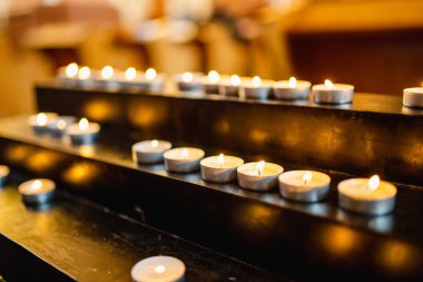 Bir dizi küçük gece lambası mumları, siyah metal bir standın üzerinde yavaşça yanar ve genellikle ibadethanelerde ya da anıtlarda bulunan barışçıl ve yansıtıcı bir ortam yaratır..