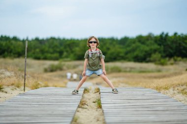 Güneş gözlüğü takan neşeli bir çocuk ve Litvanya 'daki Curonian Spit' in tahta kaldırımında neşeli bir şekilde koşan bir sırt çantası, güneşli bir açık hava macerasının tadını çıkarıyor..