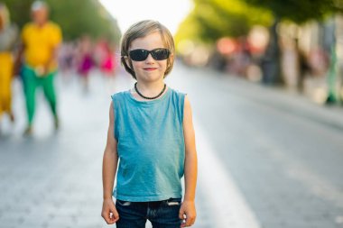 Güneşli bir günde şehrin ortasında güneş gözlüğü ve mavi atlet giyen kendine güvenen genç bir çocuk duruyor..