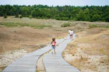 Güneş gözlüğü takan neşeli bir çocuk ve Litvanya 'daki Curonian Spit' in tahta kaldırımında neşeli bir şekilde koşan bir sırt çantası, güneşli bir açık hava macerasının tadını çıkarıyor..