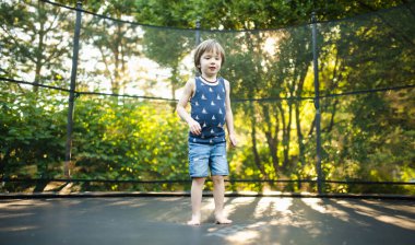 Sıcak ve güneşli bir yaz gününde, bir bahçede trambolinde zıplayan sevimli bir çocuk. Çocuklar için spor ve egzersiz. Yazın açık hava eğlence aktiviteleri.