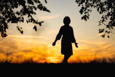 Gün batımında bir tarlada zarif bir şekilde duran bir kızın silueti, ağaçlarla çevrili, altın rengi gökyüzü, huzurlu, huzurlu bir atmosfer yaratıyor..