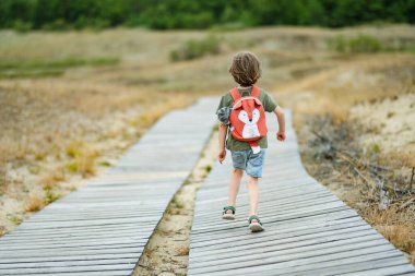 Güneş gözlüğü takan neşeli bir çocuk ve Litvanya 'daki Curonian Spit' in tahta kaldırımında neşeli bir şekilde koşan bir sırt çantası, güneşli bir açık hava macerasının tadını çıkarıyor..