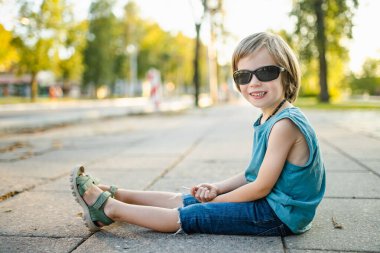 Güneş gözlüğü takan ve mavi atlet giyen gülümseyen bir çocuk güneşli bir kaldırımda öylesine oturur ve sıcak bir yaz gününün tadını çıkarır..