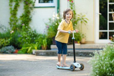 Güneşli bir yaz akşamında arka bahçede scooterını süren sevimli küçük bir çocuk. Paten süren küçük bir çocuk. Çocuklar için aktif eğlence ve açık hava sporları.