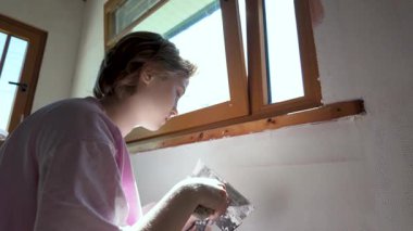 Genç kız macunu duvarın yüzeyine saçıyor. Çatlakları alçıyla doldurmak, duvarları boyamaya hazırlamak. Ev tadilatında ebeveynlere yardım ediyorum. Yavaş çekim, güneşli, parlak kapalı mekan görüntüleri..