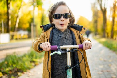 Güneşli bir sonbahar akşamında küçük sevimli bir çocuk mobiletini güneşli bir şehirde sürüyor. Paten süren küçük bir çocuk. Çocuklar için aktif eğlence ve açık hava sporları.