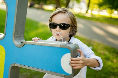 Güneş gözlüklü şık bir genç çocuk Litvanya 'nın Vilnius kentindeki güneşli bir parkta modern bir su çeşmesinden su içiyor.