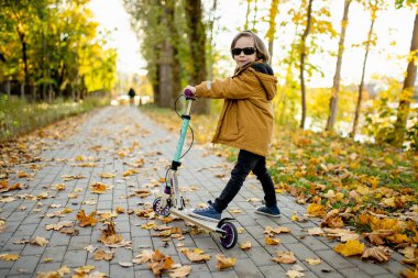 Güneşli bir sonbahar akşamında küçük sevimli bir çocuk mobiletini güneşli bir şehirde sürüyor. Paten süren küçük bir çocuk. Çocuklar için aktif eğlence ve açık hava sporları.