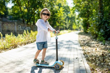 Güneşli bir yaz akşamında küçük sevimli bir çocuk mobiletiyle şehirde geziyor. Paten süren küçük bir çocuk. Çocuklar için aktif eğlence ve açık hava sporları.