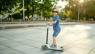 Güneşli bir yaz akşamında küçük sevimli bir çocuk mobiletiyle şehirde geziyor. Paten süren küçük bir çocuk. Çocuklar için aktif eğlence ve açık hava sporları.