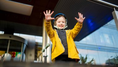 Neşeli bir çocuk heyecanlı bir şekilde iki elini kaldırır scooterlarının yanında durur, sarı ceket ve kask giyer, neşeli bir enerjiyle ışıldar.