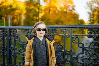 Güneş gözlüklü ve ceketli şık bir genç Vilnius 'ta bir köprüde gururla durur. Çerçevesi güz yapraklarının parlak renkleriyle süslenmiştir.
