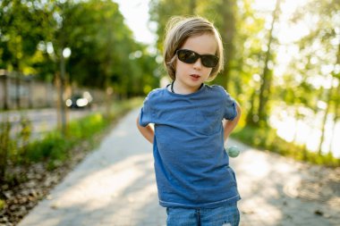 Güneş gözlüklü, mavi tişörtlü, güneşli bir kaldırımda poz veren genç çocuk.