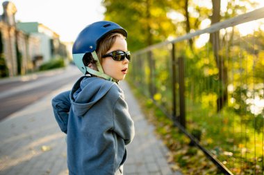 Güneşli bir sonbahar akşamında küçük sevimli bir çocuk mobiletini güneşli bir şehirde sürüyor. Paten süren küçük bir çocuk. Çocuklar için aktif eğlence ve açık hava sporları.