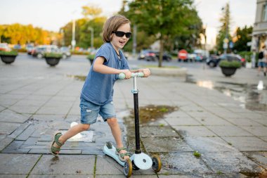 Genç bir çocuk Vilnius 'ta güneşli bir meydanda scooter sürüyor, başlık takıyor ve güneş gözlüğü takıyor, sıcak bir günün tadını çıkarıyor.