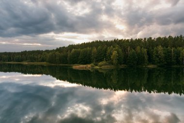 Litvanya 'daki Gela Gölü sakin sularındaki dramatik bulutları ve ormanı yansıtarak sakin ve karamsar bir manzara yaratır.