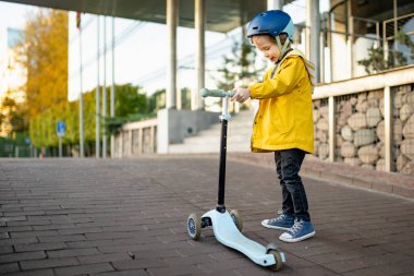 Güneşli bir sonbahar akşamında küçük sevimli bir çocuk mobiletini güneşli bir şehirde sürüyor. Paten süren küçük bir çocuk. Çocuklar için aktif eğlence ve açık hava sporları.