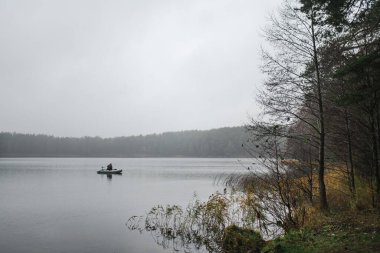 Yalnız bir insan, Balzis Gölü 'ndeki küçük bir tekneden sessizce balık tutar. Sis, sazlıklar ve yoğun ormanlarla çevrili gri gökyüzünün altında.