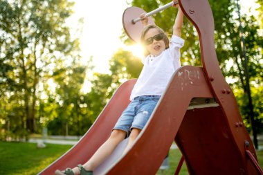 Parlak güneşli bir yaz gününde oyun alanında eğlenen sevimli küçük çocuk. Kaydırağın üzerinde sevimli bir çocuk. Çocuklu aileler için açık hava aktiviteleri.