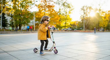 Güneşli bir sonbahar akşamında küçük sevimli bir çocuk mobiletini güneşli bir şehirde sürüyor. Paten süren küçük bir çocuk. Çocuklar için aktif eğlence ve açık hava sporları.