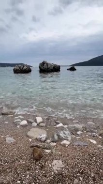 Adriyatik Denizi 'nin kristal berrak dalgaları kayalık kayalıklar ve parlak Karadağ güneşinin altında yemyeşil çam ağaçları tarafından çerçevelenmiş renkli çakıl taşlarının üzerinde yavaşça süzülüyor.