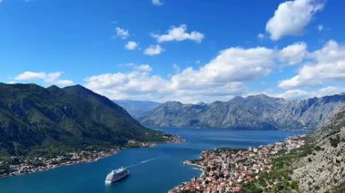 Karadağ 'ın Kotor Koyu' ndan geçen bir yolcu gemisi, görkemli dağlar, derin mavi sular ve canlı bir gökyüzü altındaki tarihi kıyı kasabalarıyla çevrili..