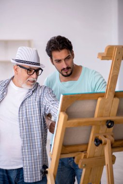 Yaşlı erkek ressam, genç erkek öğrenciye stüdyoda ders veriyor.