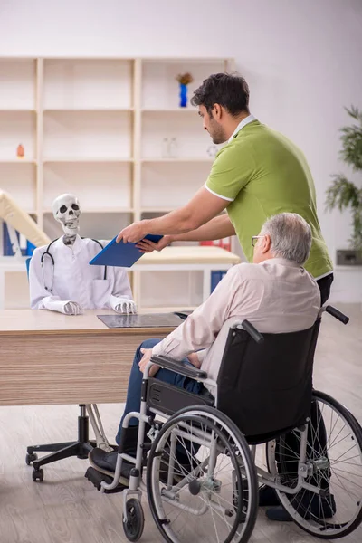 Old male patient in wheel-chair visiting skeleton doctor