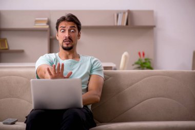 Genç bir erkek öğrenci evde bilgisayarla kanepede oturuyor.