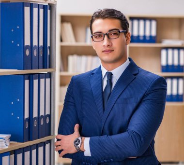 Elinde dosyalarla rafın yanında duran genç adam.