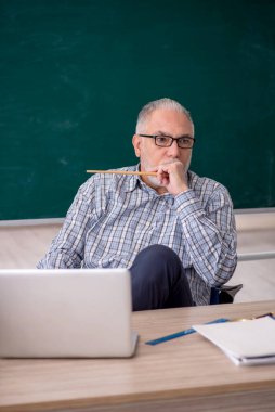 Yaşlı öğretmen yeşil tahtanın önünde oturuyor.