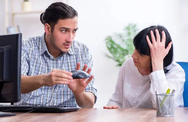 Young male employee explaining to old female colleague how to use computer