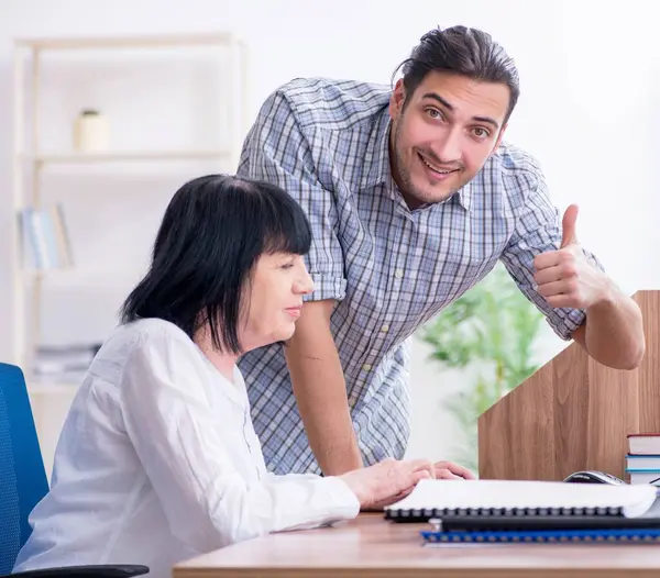 Young male employee explaining to old female colleague how to use computer