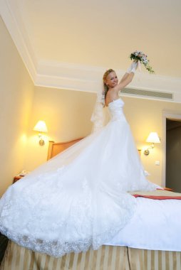 Gelin, otel odasındaki buketini kaldırarak yatakta duruyor..