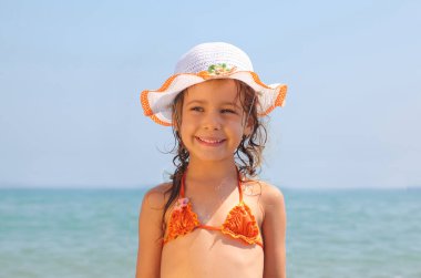 Güzel küçük bir kız, mayo ve şapka giymiş sahilde duruyor..