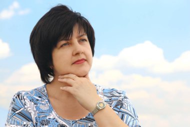 Esmer, tombul, olgun, ciddi bir kadının portresi. El çenede, mavi gökyüzü ve bulutlar.