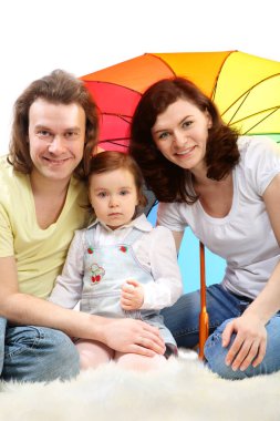 Mutlu genç aile - anne, baba ve küçük kız - beyaz tüylü bir kürkün üzerinde renkli şemsiye altında oturuyorlar.