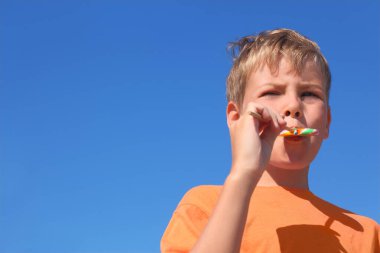 Turuncu gömlekli küçük çocuk çok renkli lolipop yiyor, mavi gökyüzü
